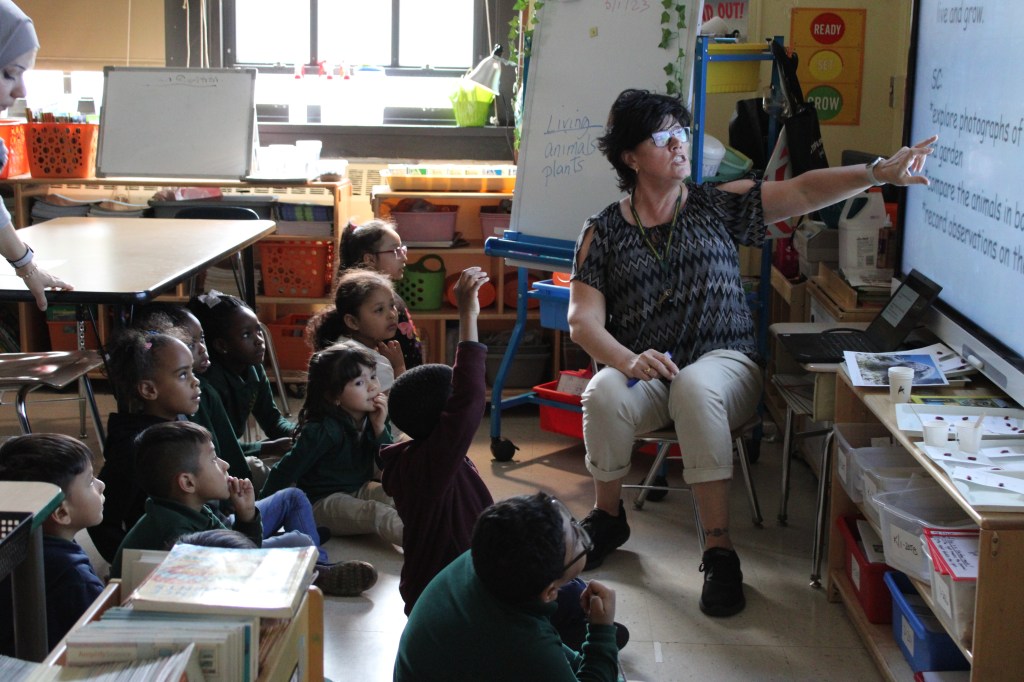 Students discuss their learning objectives with Ms. Kelly. Their teacher, Miss Kelly, is pointing to the SmartBoard