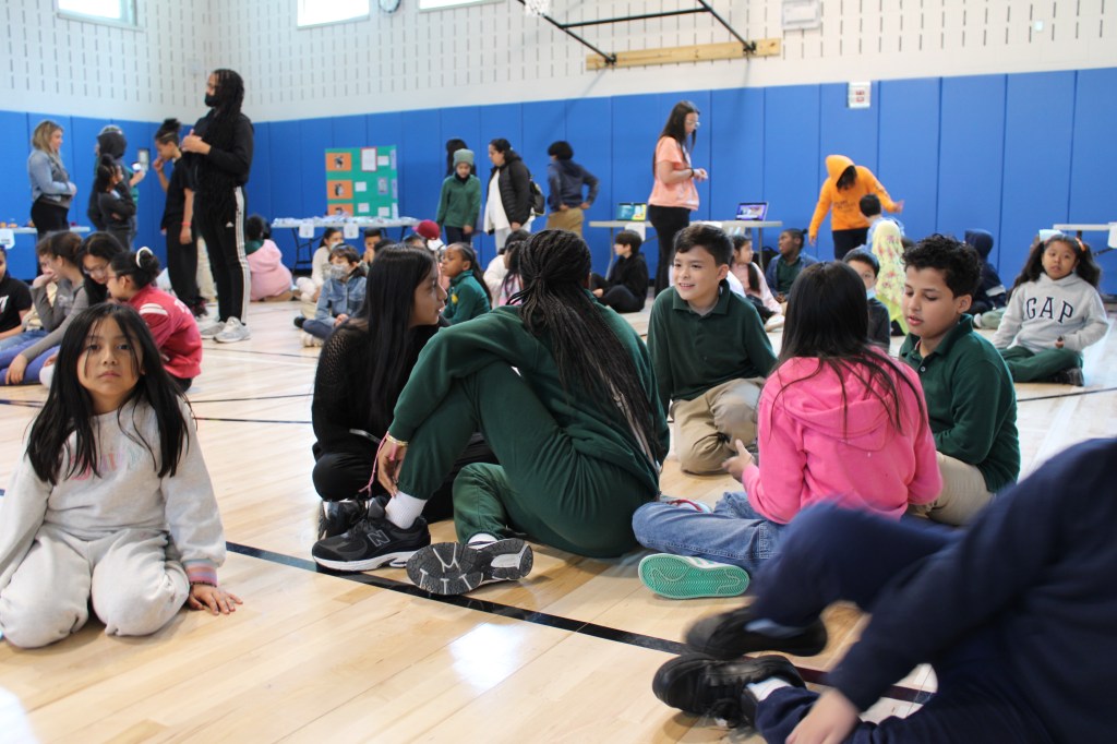 Students are sitting on the floor of the gymnasium.