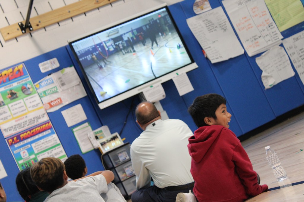 Students and teachers look at the screen during a video presentation. One student is looking back and smiling.