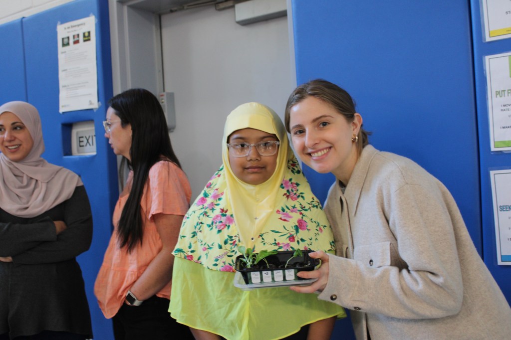 A student wearing a yellow headcovering and a teacher holding a small tray of plants pose together for a photo.