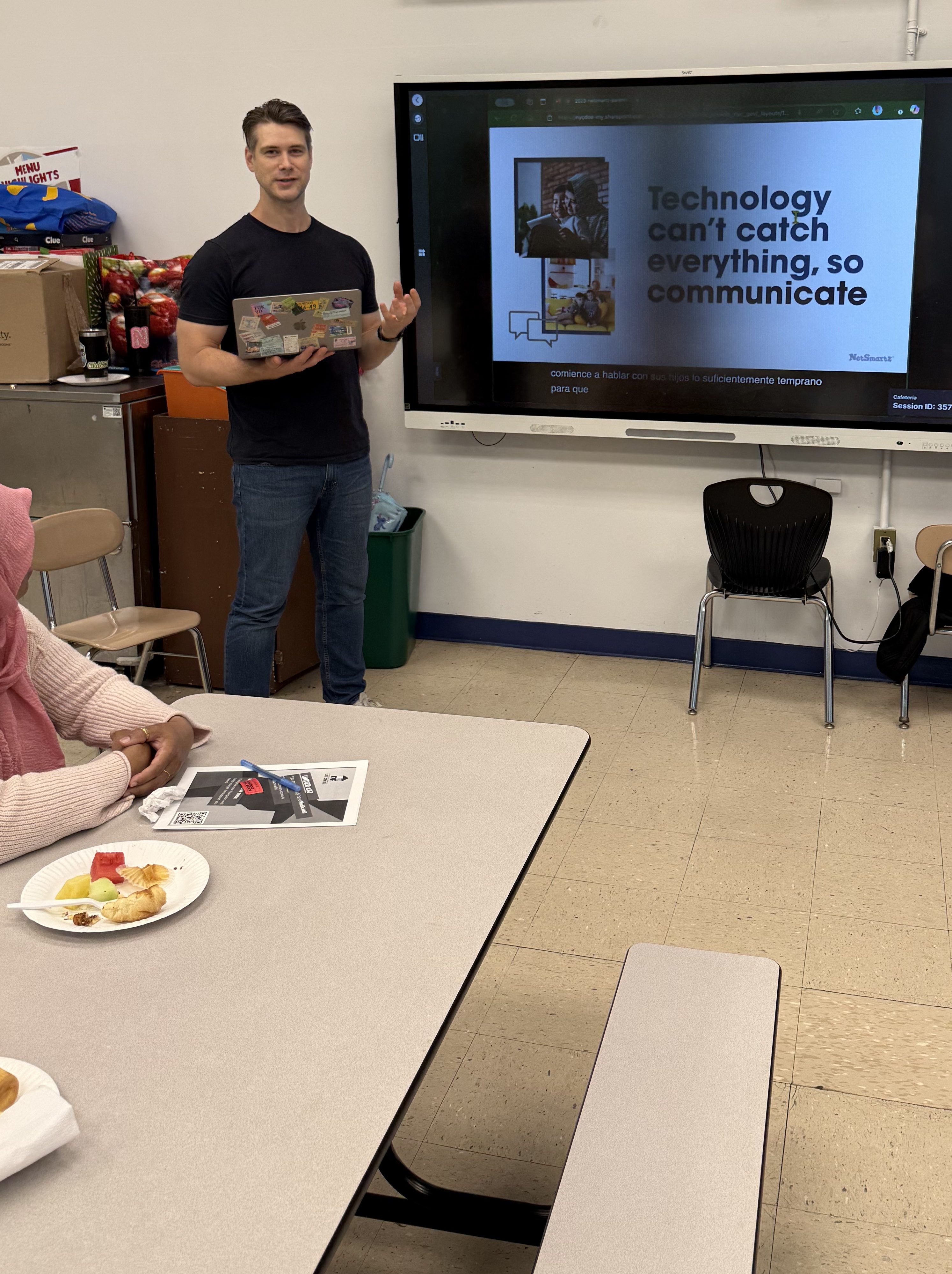 A presenter stands in front of a screen displaying a slide about technology and communication, while a participant sits at a table with food and a handout.