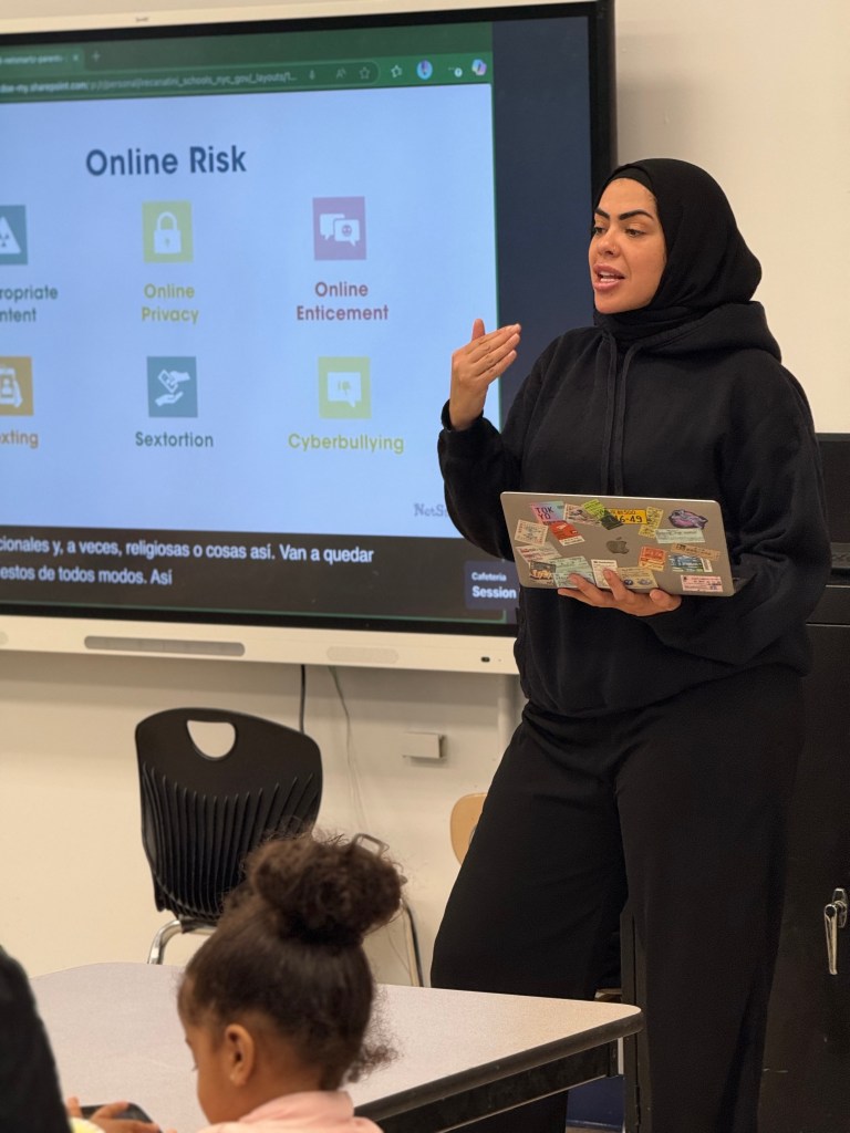 Ms. Mohamed presenting on online risks, highlighting topics like online privacy, inappropriate content, sextortion, and cyberbullying, with students seated at a table in a school setting.
