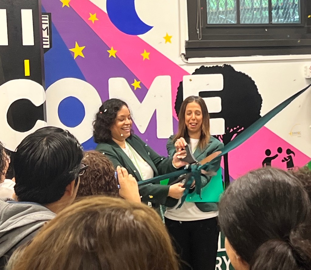 Principal Mazzeo and assistant principal La Ruffa cutting a ribbon during the ceremony in front of a colorful mural that says 'Welcome', with an audience watching.